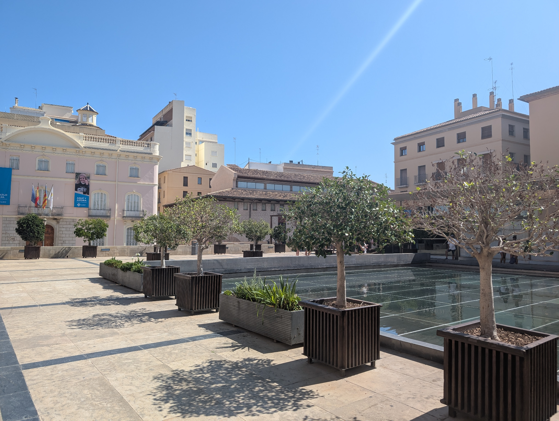 Een rustig binnenplein in Valencia met sinaasappelboompjes, een spiegelend waterbassin en pastelkleurige gevels onder een diepblauwe hemel